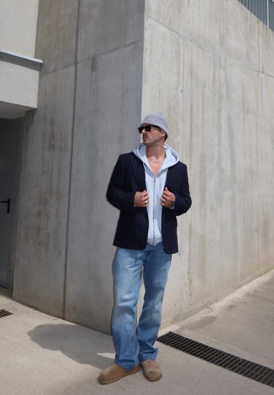 Hombre con gorro, gafas de sol, blazer negro sobre sudadera blanca con capucha, vaqueros azules y zapatos marrones, de pie junto a una pared de hormigón al aire libre.