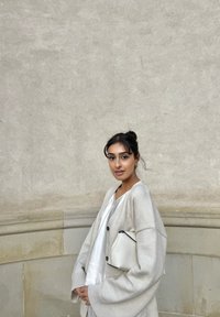 Cardigan gris clair avec des épaules tombantes, boutons en bois ; top blanc, tenant une pochette texturée blanche. Fond lisse de mur gris.