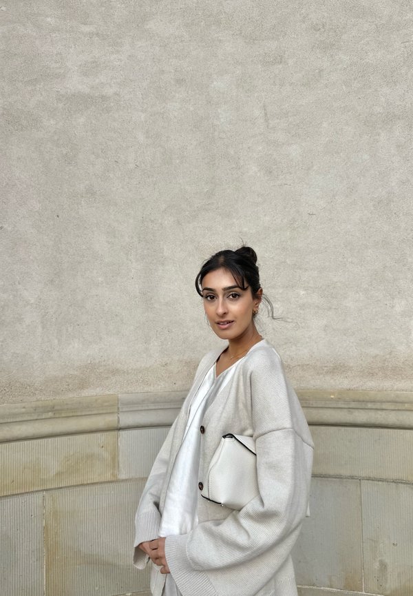 Light gray cardigan with dropped shoulders, wooden buttons; white top, holding a white textured clutch. Smooth gray wall background.