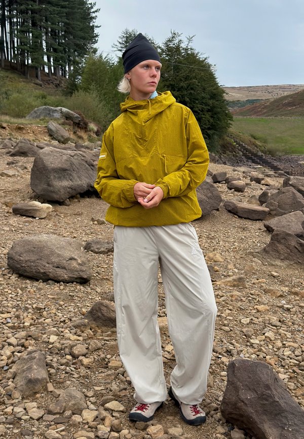 Personne portant une veste moutarde, un pantalon clair et un bonnet noir, debout sur un sol rocheux avec des arbres et des collines en arrière-plan.