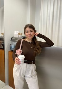 Maglione marrone a maniche lunghe, pantaloni bianchi a vita alta, cintura marrone, borsa a fiori rosa, mentre tiene una tazza di caffè, con una macchina da caffè sullo sfondo.