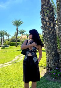 Top halter nero e bianco con motivo floreale e fronte annodato, abbinato a pantaloncini neri a vita alta e a una piccola borsa nera. Sfondo di un paesaggio verde.