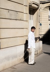 Camicia bianca oversize, pantaloni a gamba larga color crema e scarpe scure. Tiene una piccola borsa frangiata. Ambientazione urbana soleggiata.