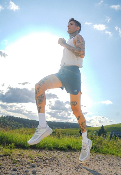 Zapatillas deportivas blancas con acentos grises, pantalones cortos ligeros y una camiseta sin mangas. La persona tiene tatuajes en los brazos y está corriendo por un camino de tierra.