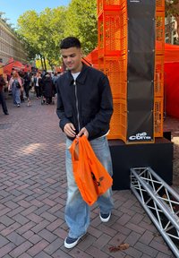 Sort zip-up jakke, hvid t-shirt, baggy lyseblå jeans og marineblå sneakers. Holder en orange tote bag. Baggrund med mursten.