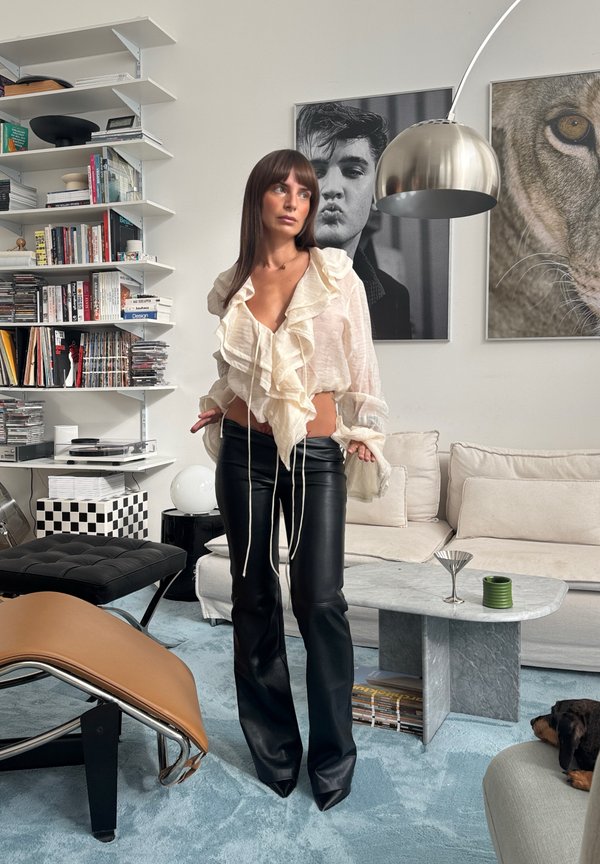 Une femme vêtue d’un chemisier blanc à volants et d’un pantalon en cuir noir se tient dans un salon moderne, à côté d’un canapé et de bibliothèques.