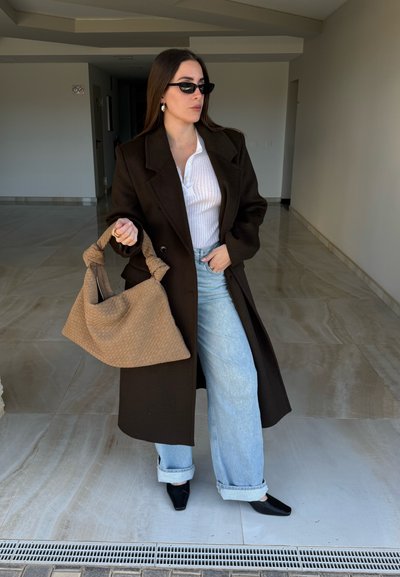 Mujer con gafas de sol negras, abrigo marrón oscuro, camiseta blanca, vaqueros azul claro y zapatos negros, sosteniendo un gran bolso beige de tejido dentro de un interior.