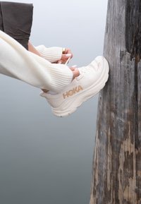 Des mains nouant les lacets d'une chaussure blanche HOKA reposant sur un poteau en bois usé à côté d'une eau calme.