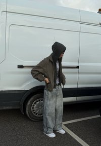 Olive roheline bomber-jakk kapuutsiga, mustriline hall särk, laiad hallid teksapüksid ja valged tossud, seismas valge kaubiku kõrval.
