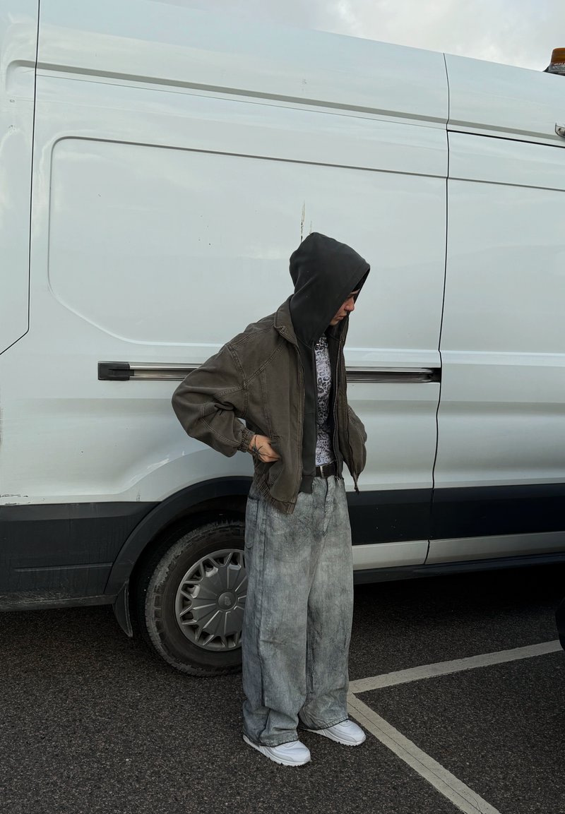 Olivgrüne Bomberjacke mit Kapuze, gemustertes graues Hemd, weite graue Jeans und weiße Sneakers, steht neben einem weißen Lieferwagen.
