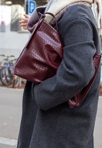 Viininpunainen kudottu nahkainen olkalaukku, pehmeä rakenne, hieman löysä muoto, pidetään harmaan villaiseen takkiin, jolla on leveät kaulukset ja hillitty huivi.