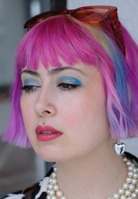 Person med en livlig pink bob-frisure, blå øjenskygge, guld næsepiercinger, hjerteformede sølvøreringe og en perlekæde.