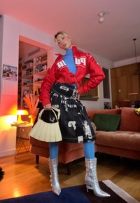 Veste rouge PUMA, haut bleu, jupe graphique noire, bottes argentées montant aux genoux, et sac à main blanc plissé ; tenue urbaine stylée aux couleurs audacieuses.