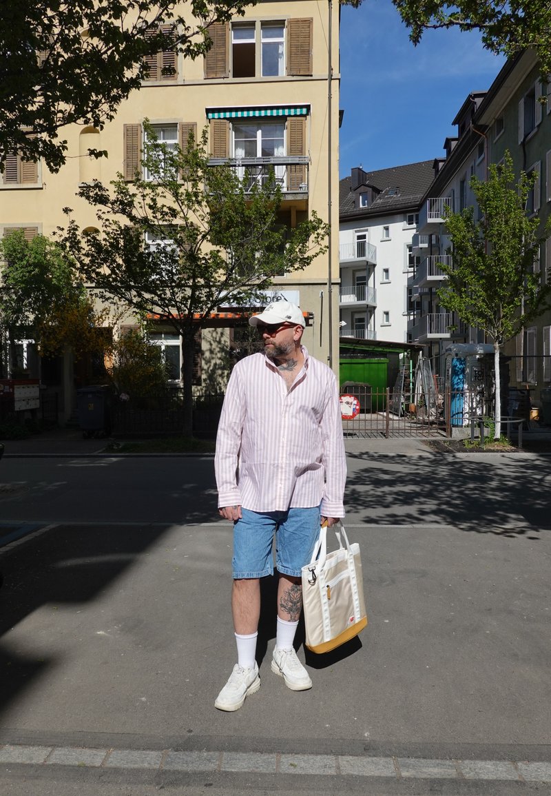 Homme barbu portant une casquette blanche, une chemise à rayures roses, un short en denim, des baskets blanches, portant un sac fourre-tout beige, debout dans une rue urbaine avec des bâtiments et des arbres.