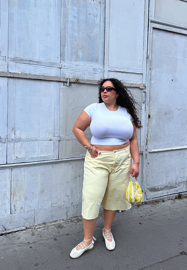 Mujer con top corto blanco y pantalones cortos anchos amarillos está apoyada contra una pared metálica gris, sostiene un bolso amarillo y lleva gafas de sol.