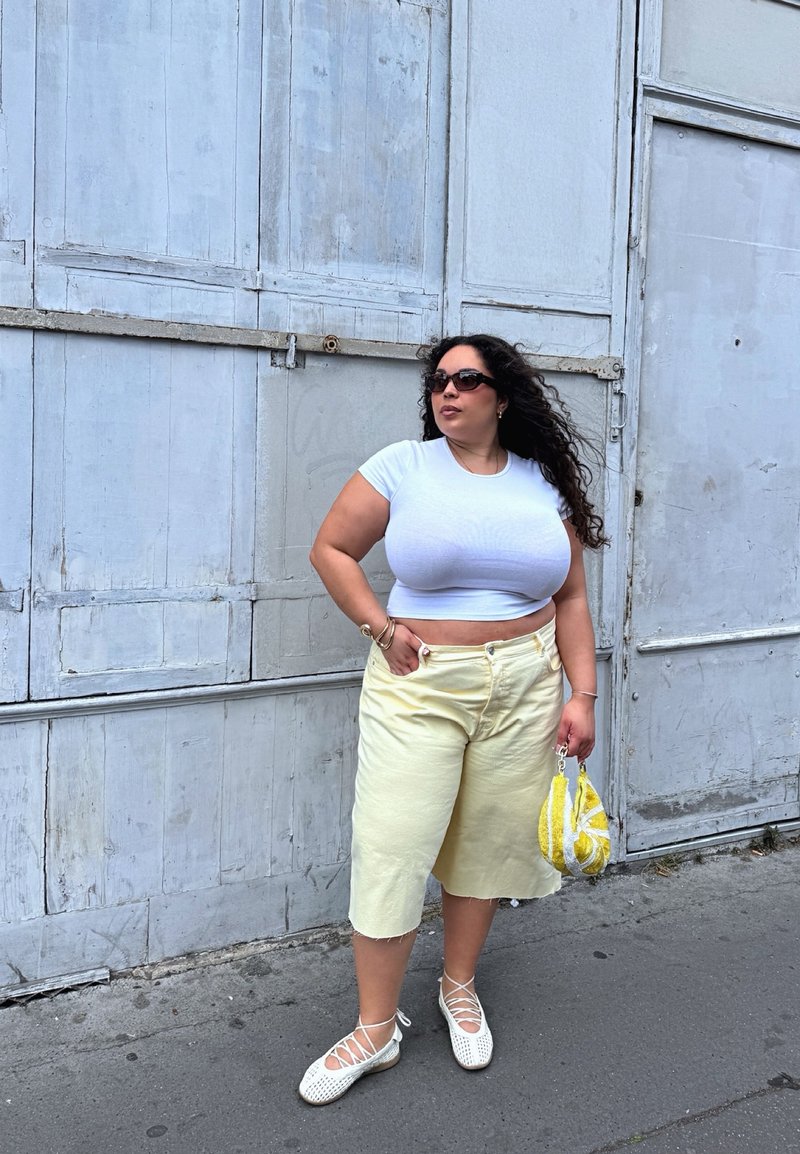 Γυναίκα φοράει λευκό crop top και κίτρινα φαρδιά σορτς, στέκεται μπροστά σε γκρι μεταλλικό τοίχο, κρατά κίτρινη τσάντα και φορά γυαλιά ηλίου.