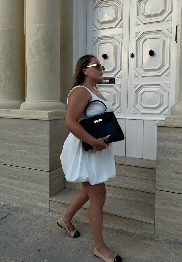 Vestito bianco senza maniche con silhouette ampia, borsa nera con dettagli dorati e scarpe beige, che cammina vicino a una grande porta bianca e colonne.