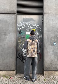 Personne portant un sweat à capuche gris, un gilet camouflage et un bonnet noir, se tenant face à une porte métallique grise couverte de graffiti sur un trottoir pavé.