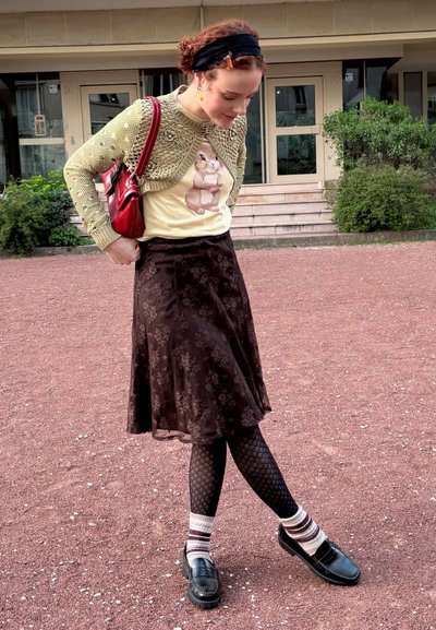 Cárdigan verde de punto con detalles decorativos sobre una camiseta amarilla con un conejo, combinado con una falda marrón con flores y medias negras estampadas.
