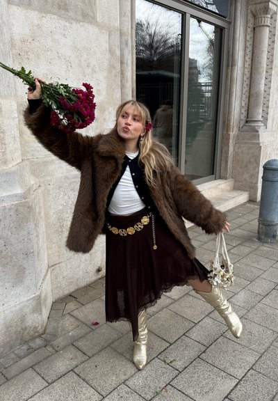 Joven mujer posando sobre una pierna en la acera de la ciudad, vestida con abrigo de piel marrón, falda negra con cinturón dorado, botas doradas, sosteniendo flores y un bolso de cuentas.