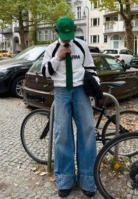 Casquette de baseball verte, chemise blanche avec manches noires, cravate verte et jean très large bleu clair. Gants noirs et baskets. Fond urbain.