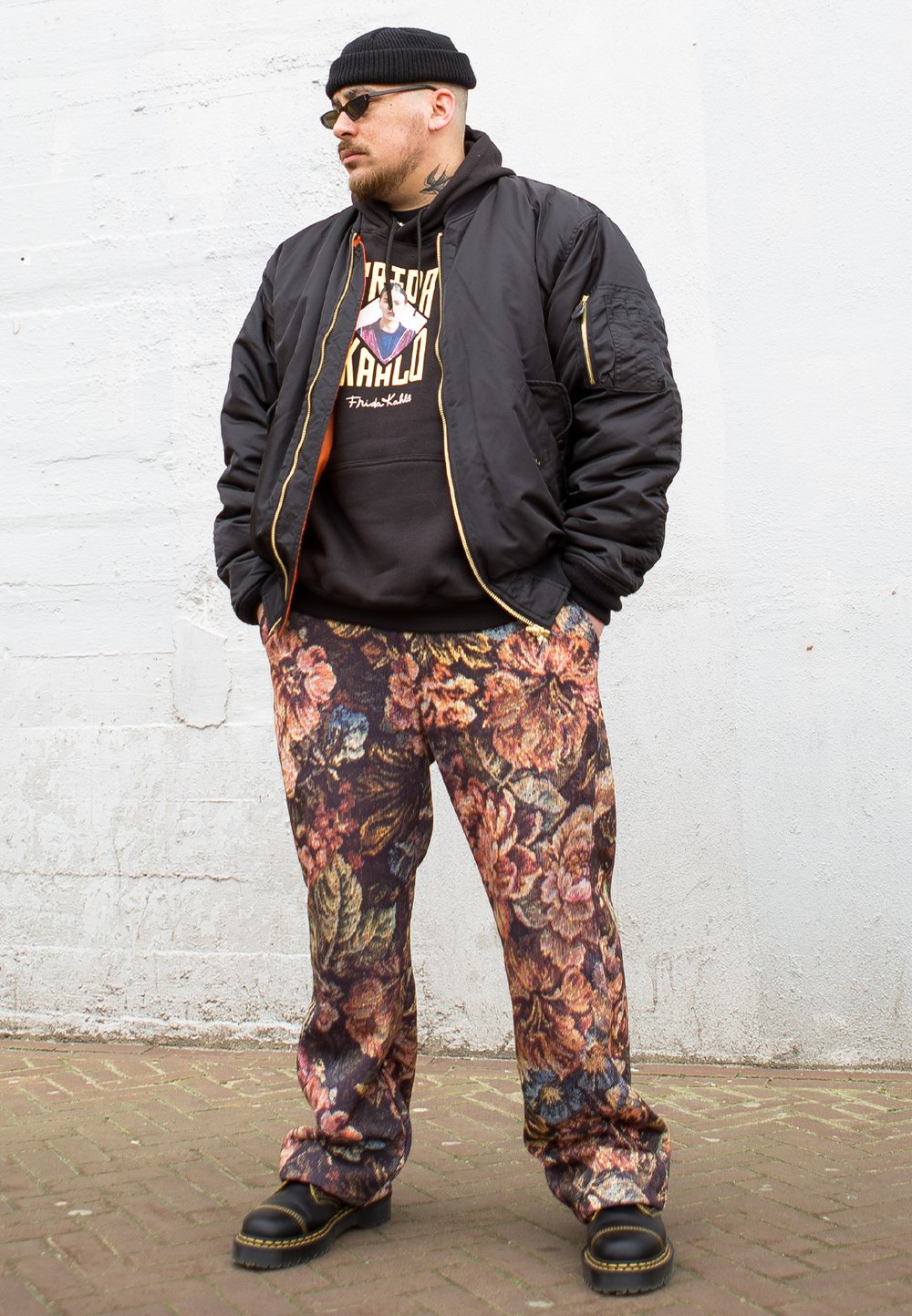 Casaco bomber preto com fechos dourados, calças com padrão floral em burgundy e dourado, hoodie preto com gráfico. Sapatos pretos robustos.