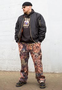Casaco bomber preto com fechos dourados, calças com padrão floral em burgundy e dourado, hoodie preto com gráfico. Sapatos pretos robustos.
