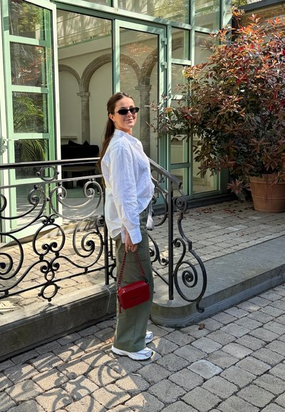 Mujer con camisa blanca y gafas de sol, sosteniendo un bolso rojo, de pie sobre un pavimento de piedra cerca de una barandilla de hierro forjado frente a un edificio de vidrio y piedra.