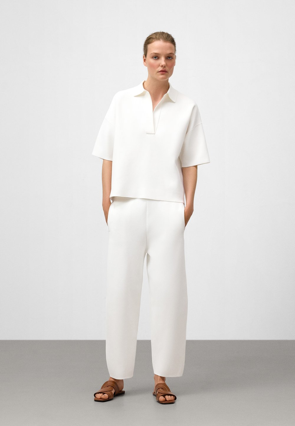 Maglietta bianca a maniche corte, morbida e con colletto, abbinata a pantaloni bianchi coordinati e sandali marroni. Tessuto liscio, design moderno.