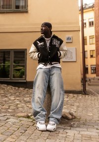 Homme portant une veste universitaire noire et blanche, un jean clair, des baskets blanches et un foulard noir, debout sur une rue pavée près de bâtiments jaunes.