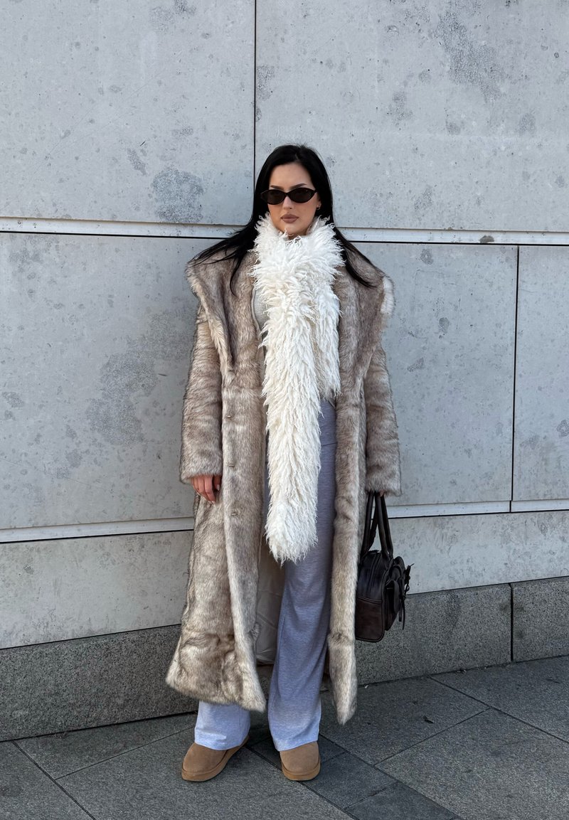 Manteau en fausse fourrure gris clair, long avec un large col. Écharpe blanche duveteuse, pantalon gris, chaussures marron clair et sac à main marron. Lunettes de soleil simples.