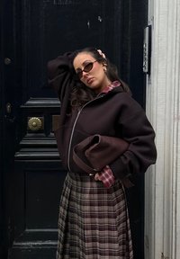 Žena v tmavé zipové bundě a kostkované sukni pózuje před černými dveřmi, drží ladící kabelku a nosí sluneční brýle a náušnice.