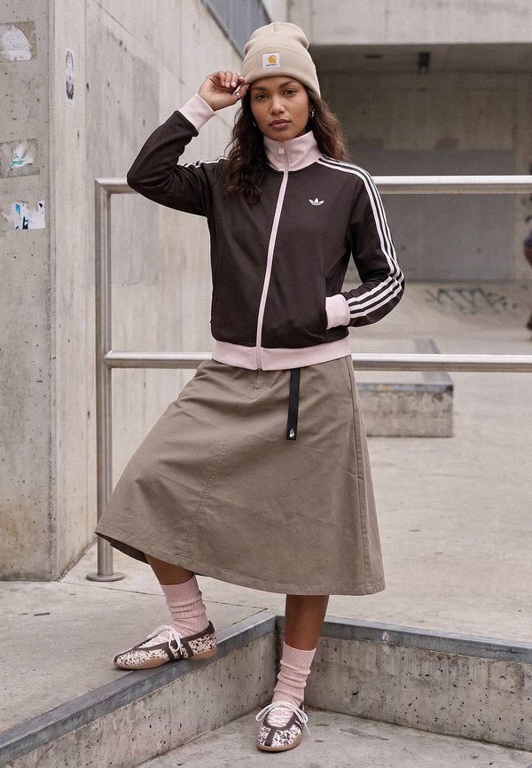 Femme portant un bonnet beige, une veste Adidas noire et rose, une jupe beige et des chaussettes roses avec des baskets à motifs, debout dans un skatepark en béton.
