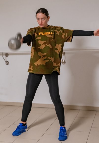 Camiseta extragrande de camuflaje con logo naranja, mangas largas negras,leggings negros y zapatillas deportivas azules. Sosteniendo un dumbbell en una pose de entrenamiento.