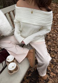 Kremasti rebrasti džemper s otkrivenim ramenima, svijetloružičaste široke hlače, smeđe gležnjače, drži dvije šalice kave u bež držaču na klupi.