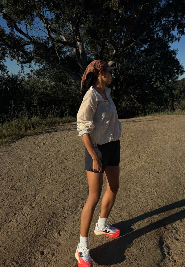 Persona con pantalones cortos, chaqueta blanca y zapatillas rojas y blancas de pie en un camino de tierra junto a árboles, mirando hacia la derecha bajo la luz del sol del final de la tarde.
