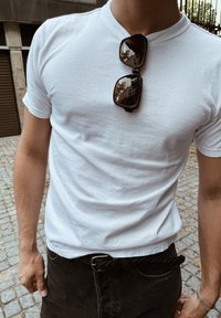 Uomo in t-shirt bianca e pantaloni neri che sta in una strada di ciottoli con occhiali da sole appesi al colletto della maglietta.
