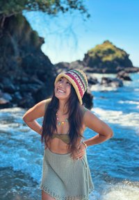 Γυναίκα με πολύχρωμο καπέλο, πλεκτή φούστα και κολιέ στέκεται χαμογελαστή κοντά σε βραχώδη ακτή με κύματα και γαλάζιο ουρανό στο φόντο.