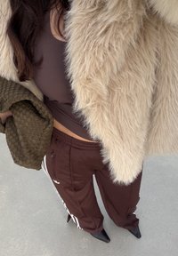 Cappotto in pelliccia sintetica marrone sopra una maglia aderente marrone, abbinato a pantaloni sportivi marroni con strisce bianche e décolleté nere con punta affilata.