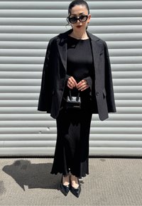 Črn tailoring blazer preko črne rebraste obleke z razširjenimi rokavi. Dodatki vključujejo majhno črno torbico in črne modne čeveljce.