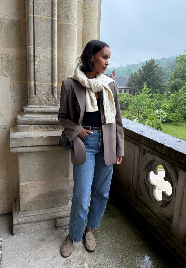 Femme portant un blazer marron, un pull blanc reposant sur les épaules, un jean bleu et des mocassins, s'appuyant contre un balcon en pierre surplombant un jardin.