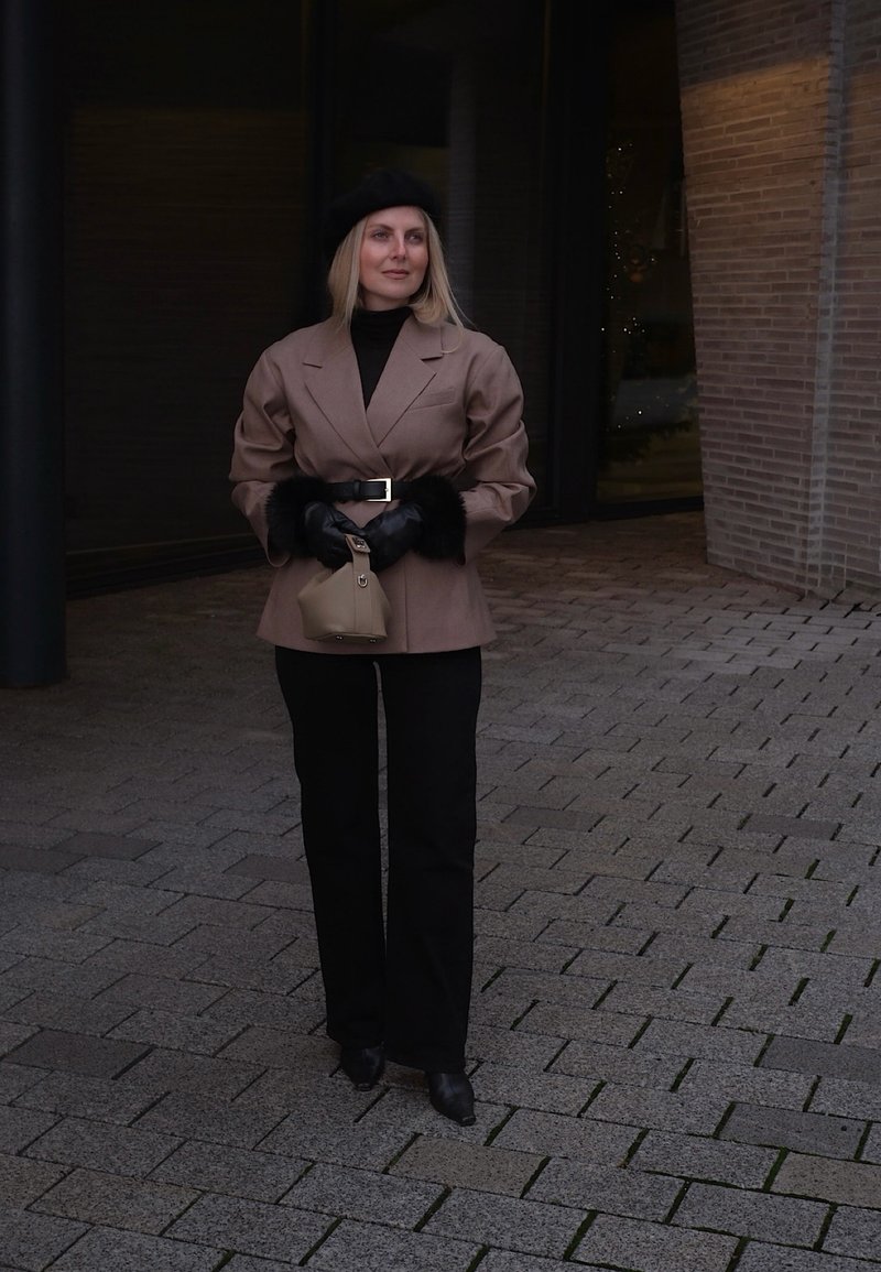 Donna che indossa un blazer marrone con cintura e polsini in pelliccia, pantaloni neri, guanti e basco, che tiene una borsa beige su una strada lastricata.