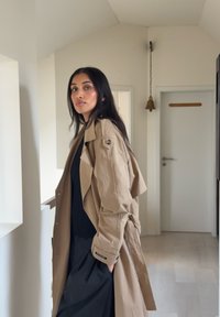 Trench-coat brun clair à coupe décontractée, avec une épaulette unique sur l'épaule, associé à un haut noir côtelé et une jupe noire fluide.
