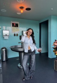 Valkoinen nappipaita, jossa on kerätyt hihansuut, yhdistettynä väljiin tummiin farkkuihin. Ruostumattomasta teräksestä valmistettu allas ja vaaleanvihreä laatta taustalla.