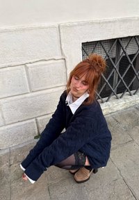 Jeune femme aux cheveux roux attachés en chignon, accroupie contre un mur en pierre blanc avec une fenêtre à barreaux, portant un pull bleu marine et des bas.