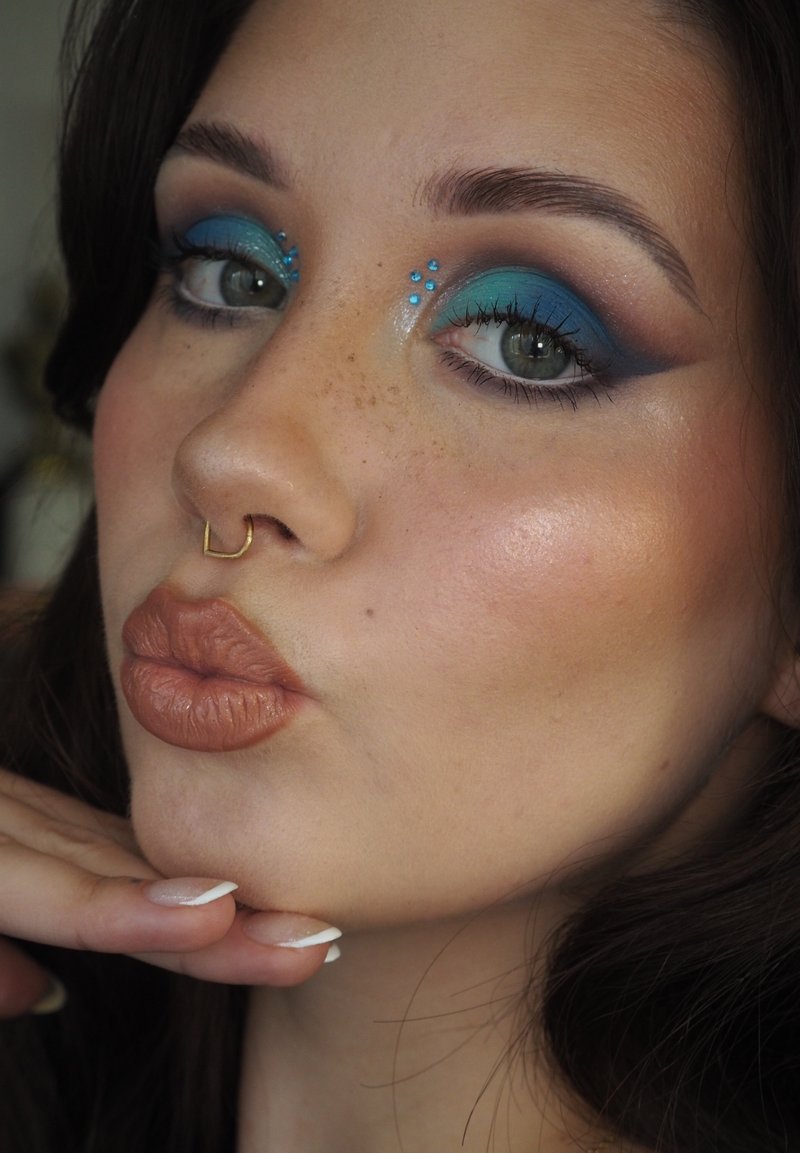 Gros plan sur un visage avec un fard à paupières bleu et des pierres précieuses sous les yeux. Les traits comprennent un piercing au septum, des lèvres pulpeuses avec un rouge à lèvres brun et des ongles manucurés.