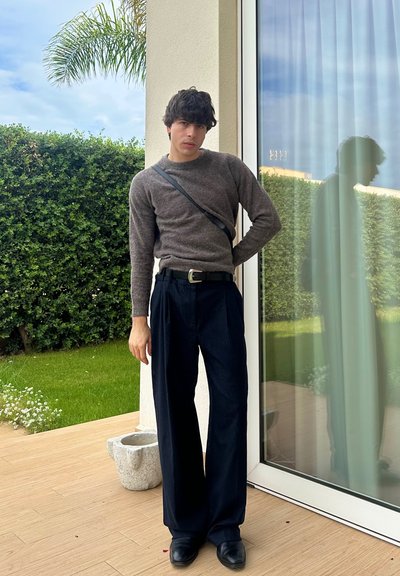 Suéter de punto gris y pantalones de pierna ancha azul marino, de pie en un patio de madera, con plantas verdes y una puerta de cristal al fondo.