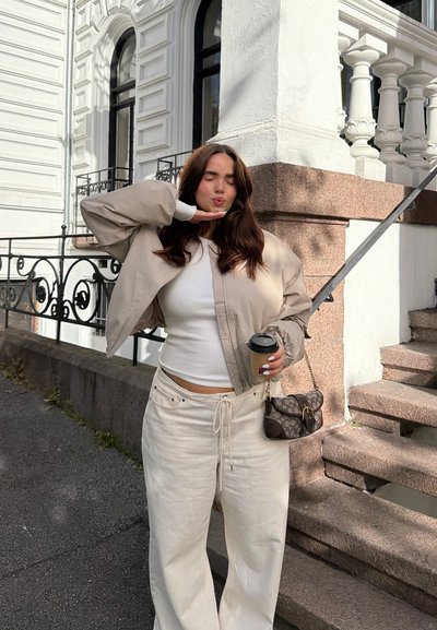 Joven mujer con chaqueta beige y pantalones blancos sosteniendo una taza de café, de pie junto a unos escalones de piedra y una barandilla de hierro fuera de un edificio blanco.