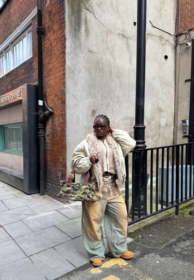 Persona con cabello trenzado que lleva una chaqueta de piel beige sobredimensionada, pantalones tie-dye, gafas de sol y sostiene un bolso de camuflaje, de pie junto a una pared urbana.