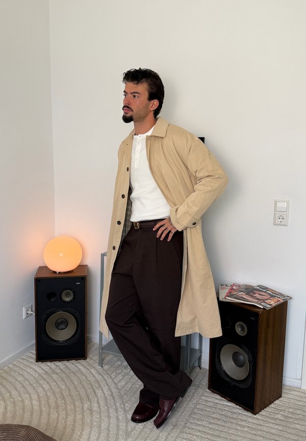 Hombre con barba, con abrigo beige, camisa blanca y pantalones oscuros, apoyado contra una pared entre dos altavoces en una habitación minimalista.